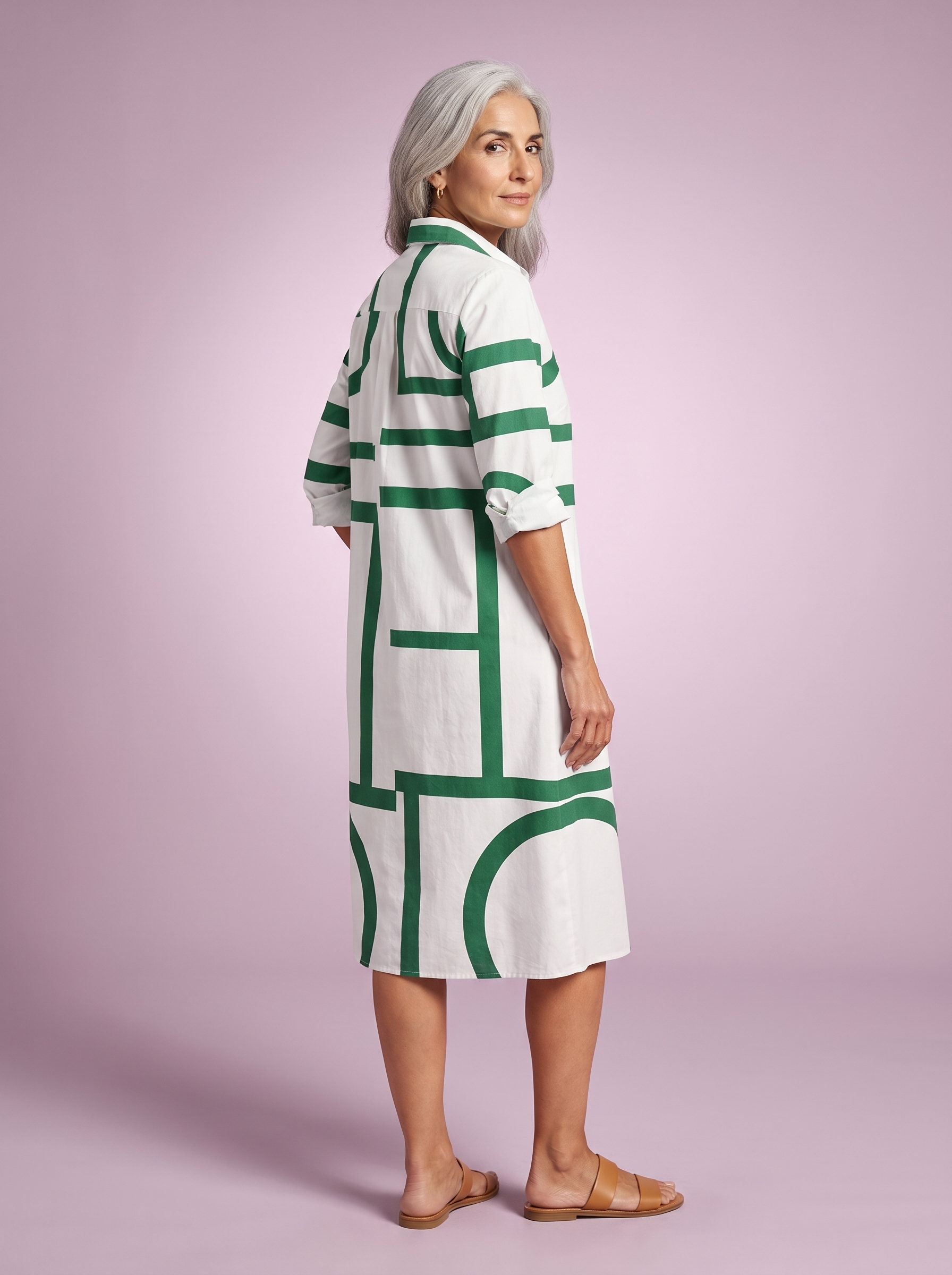 Spanish woman over 50 in the white shirt dress with a green geometric line print, three-quarter back studio view on a lilac backdrop