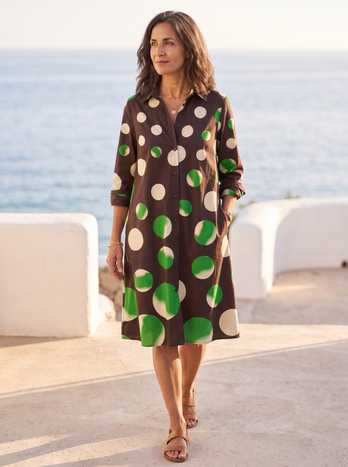 Spanish woman over 50 walking a sunlit coastal promenade in the chocolate-brown polka-dot shirt dress