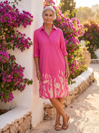 Spanish woman over 50 leaning by a whitewashed garden wall in the fuchsia botanical shirt dress